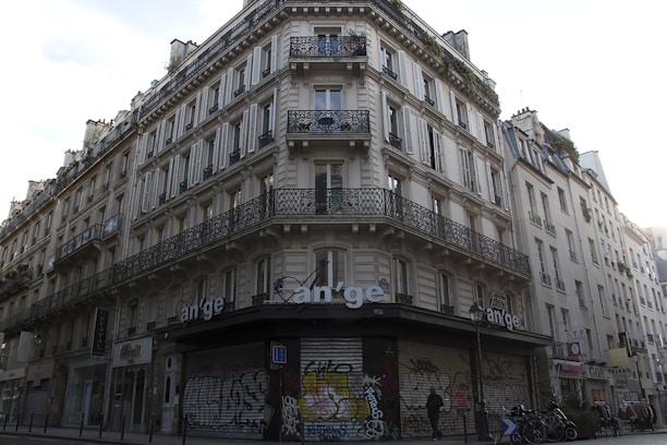 A classic Parisian building with ornate wrought iron balconies and white shutters on multiple floors. The street-level facade is covered in graffiti, and there are bicycles parked at the corner. A person is walking by, adding a sense of urban life to the scene.