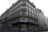 A classic Parisian building with ornate wrought iron balconies and white shutters on multiple floors. The street-level facade is covered in graffiti, and there are bicycles parked at the corner. A person is walking by, adding a sense of urban life to the scene.