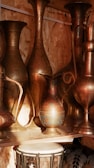 A collection of intricately designed brass vessels and jugs is displayed on a wooden shelf. The patterns on the metal surfaces are detailed and exhibit traditional craftsmanship. Warm lighting accentuates their metallic shine, creating a cozy and inviting atmosphere. Below the vessels, a traditional hand drum with a patterned surface is partially visible.