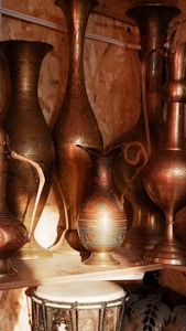 A collection of intricately designed brass vessels and jugs is displayed on a wooden shelf. The patterns on the metal surfaces are detailed and exhibit traditional craftsmanship. Warm lighting accentuates their metallic shine, creating a cozy and inviting atmosphere. Below the vessels, a traditional hand drum with a patterned surface is partially visible.