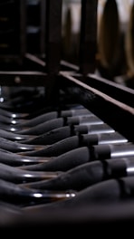 A close-up view of several dark glass wine bottles stored upside down in a wine rack, creating a repetitive pattern. The lighting is dim, highlighting the sleek contours of the bottles and the metal structure of the rack.