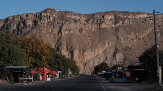 A wide road runs through a small rural area nestled at the base of a large, rocky mountain. On both sides of the road, there are small shops with signs and umbrellas, surrounded by trees. The mountainous backdrop is rugged and majestic, with a clear blue sky above.