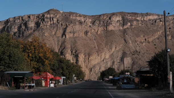 A wide road runs through a small rural area nestled at the base of a large, rocky mountain. On both sides of the road, there are small shops with signs and umbrellas, surrounded by trees. The mountainous backdrop is rugged and majestic, with a clear blue sky above.