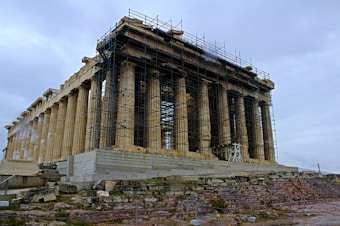 An ancient stone temple with a series of columns undergoing restoration, surrounded by scaffolding. The structure stands on a rocky, uneven terrain with a cloudy sky overhead.