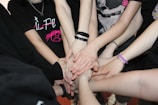 Several people stand in a circle, extending one hand each towards the center, all touching in a gesture of unity and teamwork. They wear casual clothing, with bracelets and wristbands visible on some arms.