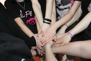 Several people stand in a circle, extending one hand each towards the center, all touching in a gesture of unity and teamwork. They wear casual clothing, with bracelets and wristbands visible on some arms.