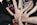 Several people stand in a circle, extending one hand each towards the center, all touching in a gesture of unity and teamwork. They wear casual clothing, with bracelets and wristbands visible on some arms.