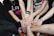 Several people stand in a circle, extending one hand each towards the center, all touching in a gesture of unity and teamwork. They wear casual clothing, with bracelets and wristbands visible on some arms.