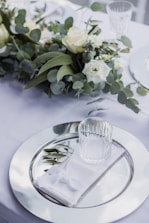A table setting with a large glass charger plate, a folded white napkin, and a clear glass resting on top. Greenery, including eucalyptus and roses, is arranged along the center of the table, creating an elegant and fresh atmosphere.