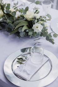 A table setting with a large glass charger plate, a folded white napkin, and a clear glass resting on top. Greenery, including eucalyptus and roses, is arranged along the center of the table, creating an elegant and fresh atmosphere.