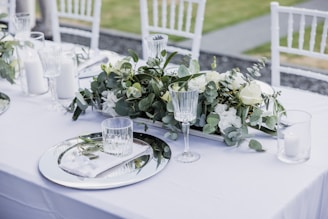 A beautifully set table with a white tablecloth, adorned with elegant glassware and plates. A lush green and white floral centerpiece runs down the length of the table, accompanied by white candles in glass holders. The chairs are white and have a classic design.