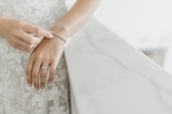 A hand wearing a wedding ring and an elegant bracelet, positioned on a marble surface with a floral lace fabric in the background.