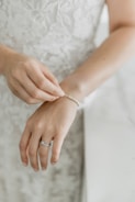 A close-up of a person's hands with one hand clasping a delicate pearl bracelet on the wrist of the other hand. The hands are adorned with a ring, and the background features a softly blurred, detailed white lace fabric.