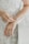 A close-up of a person's hands with one hand clasping a delicate pearl bracelet on the wrist of the other hand. The hands are adorned with a ring, and the background features a softly blurred, detailed white lace fabric.