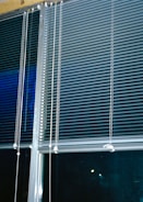 Horizontal blinds cover three large window panels. The blinds are partially closed, with several cords hanging down to adjust their position. The background outside the window is dark, suggesting it might be nighttime.
