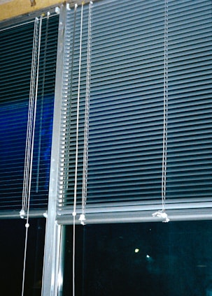 Horizontal blinds cover three large window panels. The blinds are partially closed, with several cords hanging down to adjust their position. The background outside the window is dark, suggesting it might be nighttime.