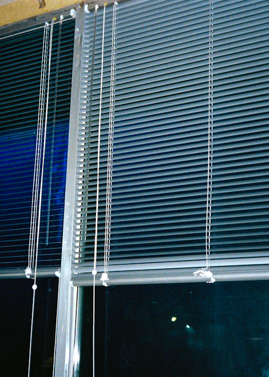 Horizontal blinds cover three large window panels. The blinds are partially closed, with several cords hanging down to adjust their position. The background outside the window is dark, suggesting it might be nighttime.