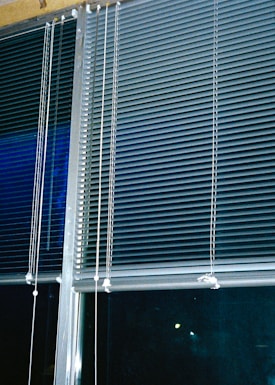 Horizontal blinds cover three large window panels. The blinds are partially closed, with several cords hanging down to adjust their position. The background outside the window is dark, suggesting it might be nighttime.