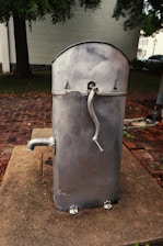 A vintage, metallic hand-cranked water pump stands on a concrete base in an outdoor setting. The pump has a curved top with a spout extending from its side and sits on a brick-paved ground surrounded by trees and a building in the background.
