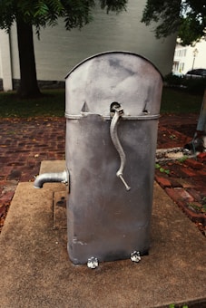 A vintage, metallic hand-cranked water pump stands on a concrete base in an outdoor setting. The pump has a curved top with a spout extending from its side and sits on a brick-paved ground surrounded by trees and a building in the background.