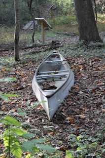 A canoe is situated on the ground amidst a wooded area with fallen leaves and greenery surrounding it. Nearby, wooden signposts are visible, suggesting a nature trail or park setting. The canoe appears unused and blends into the natural environment.