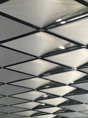 A geometric pattern of triangular fabric panels supported by a framework of black metal beams. The white panels are arranged in a repetitive, crisscrossing design. Light filtering from above creates a subtle play of shadows and highlights on the fabric.