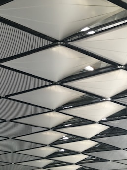 A geometric pattern of triangular fabric panels supported by a framework of black metal beams. The white panels are arranged in a repetitive, crisscrossing design. Light filtering from above creates a subtle play of shadows and highlights on the fabric.
