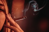 A close-up of a person holding a lit cigar between their fingers, with smoke curling upwards. The lighting is warm and dim, creating a moody atmosphere.