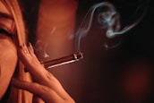 A close-up of a person holding a lit cigar between their fingers, with smoke curling upwards. The lighting is warm and dim, creating a moody atmosphere.