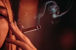 A close-up of a person holding a lit cigar between their fingers, with smoke curling upwards. The lighting is warm and dim, creating a moody atmosphere.