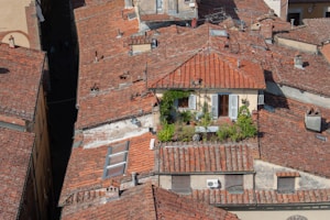 An overhead view of a cluster of historic buildings featuring red-tiled roofs. A rooftop garden with plants and greenery can be seen on one roof, indicating a residential space. The setting appears to be a dense, urban area with tightly packed buildings.