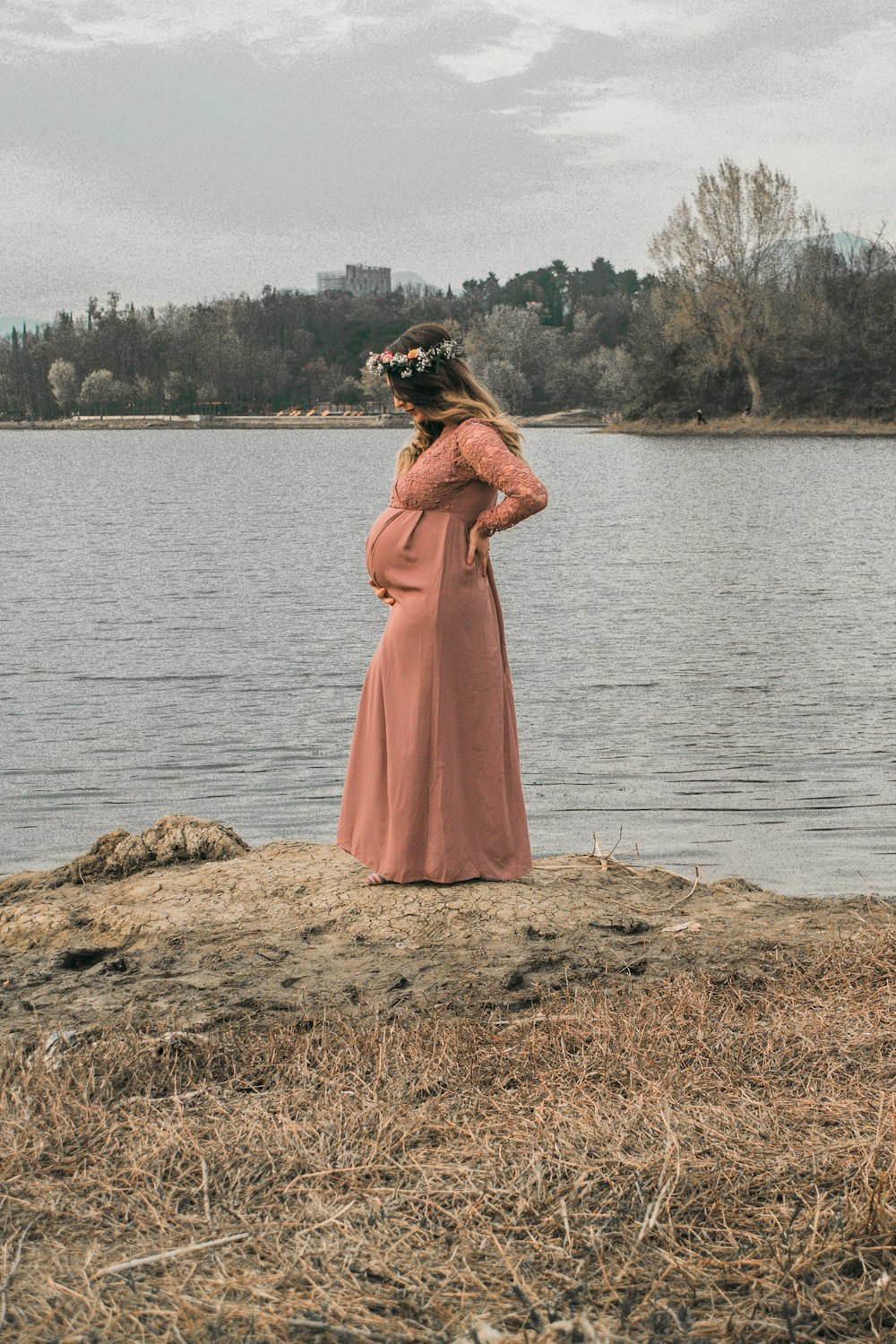 a pregnant woman in a pink dress standing on a rock by a lake