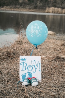 A blue balloon with the phrase 'It's a Boy!' is tethered to a card displaying the same message and decorated with footprints. Below the card is a pair of small blue baby shoes placed on dried grass beside a calm body of water, creating a rustic aesthetic.