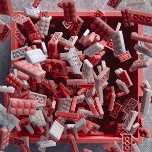 A collection of interlocking brick toys in red and white colors is falling into a square container with an open top. The bricks are captured mid-motion against a soft-focused, textured background.