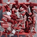 A collection of interlocking brick toys in red and white colors is falling into a square container with an open top. The bricks are captured mid-motion against a soft-focused, textured background.