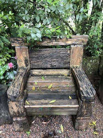A rustic wooden bench surrounded by lush green foliage. The bench appears weathered and aged, with wet leaves scattered across its seat. The setting is natural, with a dense arrangement of plants and trees in the background, creating a tranquil forest-like atmosphere.