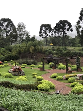 A lush garden features neatly trimmed shrubs and green grass, with a winding gravel path leading to a pergola. A rustic wooden cart filled with flowers is prominently placed, and tall trees line the background under a cloudy sky. A bright yellow umbrella stands out in the middle of the garden.