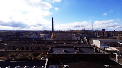 A sprawling industrial complex with multiple factory buildings and smokestacks, under a partly cloudy sky. The landscape is mostly flat with a few tall structures like communication towers in the background.