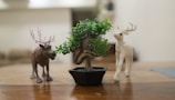 A small artificial bonsai tree in a black pot is placed on a wooden surface, flanked by two toy reindeer figurines on either side. The background is blurred, suggesting an indoor setting with neutral colors.
