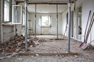 A partially demolished room with debris scattered across the floor. There are open windows along the left wall and upright metallic support beams in the center. A ladder leans against the back wall, and various construction tools are visible along the right wall.
