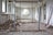 A partially demolished room with debris scattered across the floor. There are open windows along the left wall and upright metallic support beams in the center. A ladder leans against the back wall, and various construction tools are visible along the right wall.