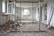 A partially demolished room with debris scattered across the floor. There are open windows along the left wall and upright metallic support beams in the center. A ladder leans against the back wall, and various construction tools are visible along the right wall.