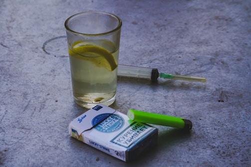 A glass filled with a light beverage contains a lemon slice, set on a concrete surface. Nearby, a syringe with a green cap, a green lighter, and a pack of cigarettes are placed casually around.