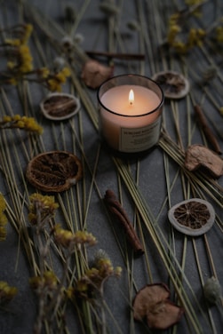 A lit candle placed among dried botanical elements, including slices of dried citrus fruits, cinnamon sticks, and yellow dried flowers. These objects are arranged on a dark, textured surface, creating a calm and rustic ambiance.