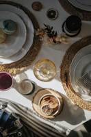 An elegantly set table features layered plates on woven placemats, with a variety of small decorative items. Macarons are placed on one of the plates, and a small dry floral arrangement adds a natural touch to the setting. There are also candles and a bowl with dried citrus slices, suggesting a cozy, rustic ambiance.