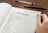 A hand holding open a book showing a table of contents, listing chapters and sections with their corresponding page numbers. Two pens are placed on the wooden surface beside the book.