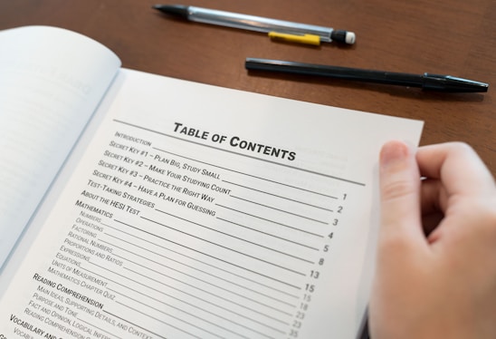 A hand holding open a book showing a table of contents, listing chapters and sections with their corresponding page numbers. Two pens are placed on the wooden surface beside the book.