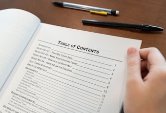 A hand holding open a book showing a table of contents, listing chapters and sections with their corresponding page numbers. Two pens are placed on the wooden surface beside the book.