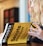 A person with long, wavy hair is holding a book titled 'Mometrix Test Preparation: Secrets Study Guide'. The cover is bright yellow with black text. The background appears blurred, focusing on the book.