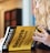 A person with long, wavy hair is holding a book titled 'Mometrix Test Preparation: Secrets Study Guide'. The cover is bright yellow with black text. The background appears blurred, focusing on the book.